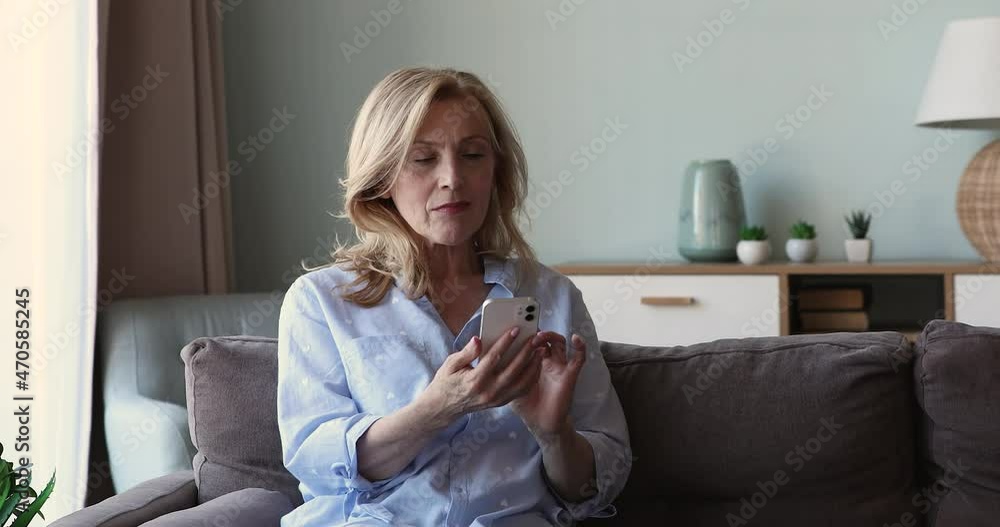 Senior people and tech. Focused aged woman sit on sofa at living room write message in chat watch video browse internet on cellphone screen. Older lady relax at home using smartphone networking in app Senior people and tech. Focused aged woman sit on sofa at living room write message in chat watch video browse internet on cellphone screen. Older lady relax at home using smartphone networking in app