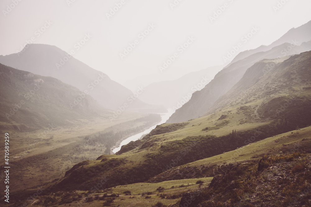 Vintage misty landscape with hills and rocks on background of wide mountain river in mist in sepia tones. Gloomy scenery with mountain relief and big river in valley in rainy weather in faded colors.