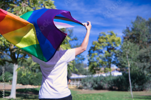 Non-binary person, with his back turned, raising with his hands the new lgbtq pride flag that is waving in the wind. Non-binary concept, lgbtq+, inclusion, gay, lesbian, lgbtq.