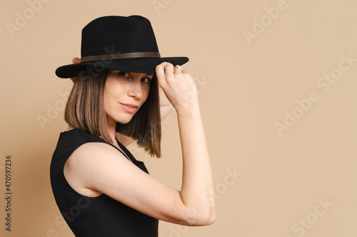 Elegance portrait of a young stylish woman in a black hat. Girl posing on beige background, studio. Beautiful girl wearing classic black dress.