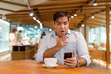 Shocked and surprised Persian man sitting at coffee shop while using mobile phone