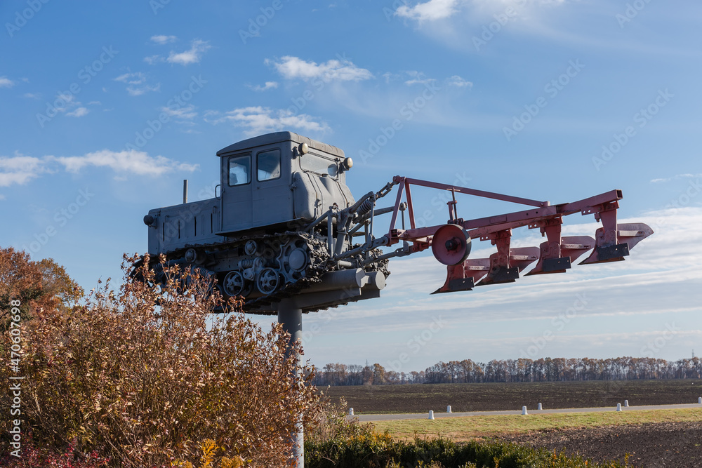 Fototapeta premium Monument to old caterpillar tractor with mounted plough against field