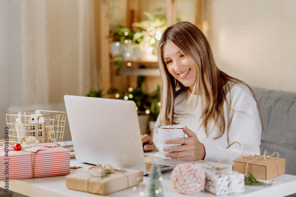 Happy and smiling young woman wrapping and preparing Christmas gifts, typing at laptop and having online video chat. Online shopping at Christmas holidays. Home interior with lights and plants Happy and smiling young woman wrapping and preparing Christmas gifts, typing at laptop and having online video chat. Online shopping at Christmas holidays. Home interior with lights and plants