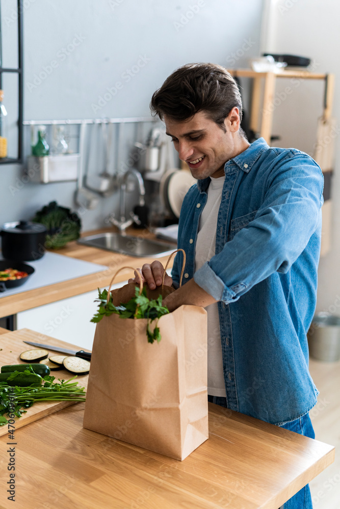 Smiling man removing vegetables from shopping bag in kitchen Smiling man removing vegetables from shopping bag in kitchen