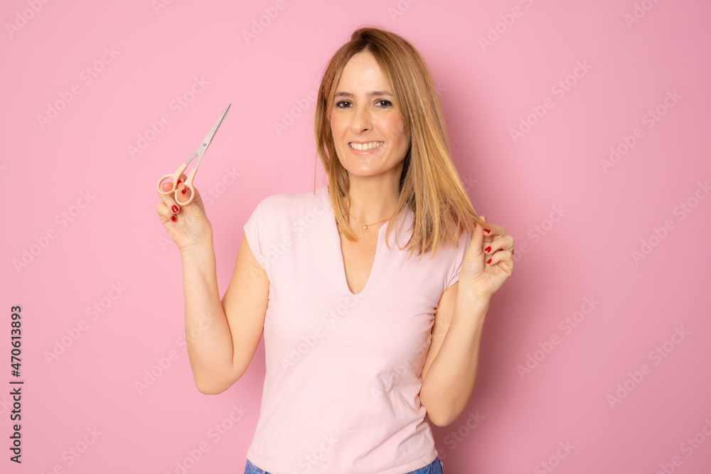 Young beautiful woman cutting her hair tips with scissors isolated over pink background.