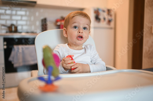 Little happy cute baby toddler boy blonde sitting on baby chair playing with constructor. Baby facial expressions indoors at home kitchen interior with toys. Healthy happy family childhood concept