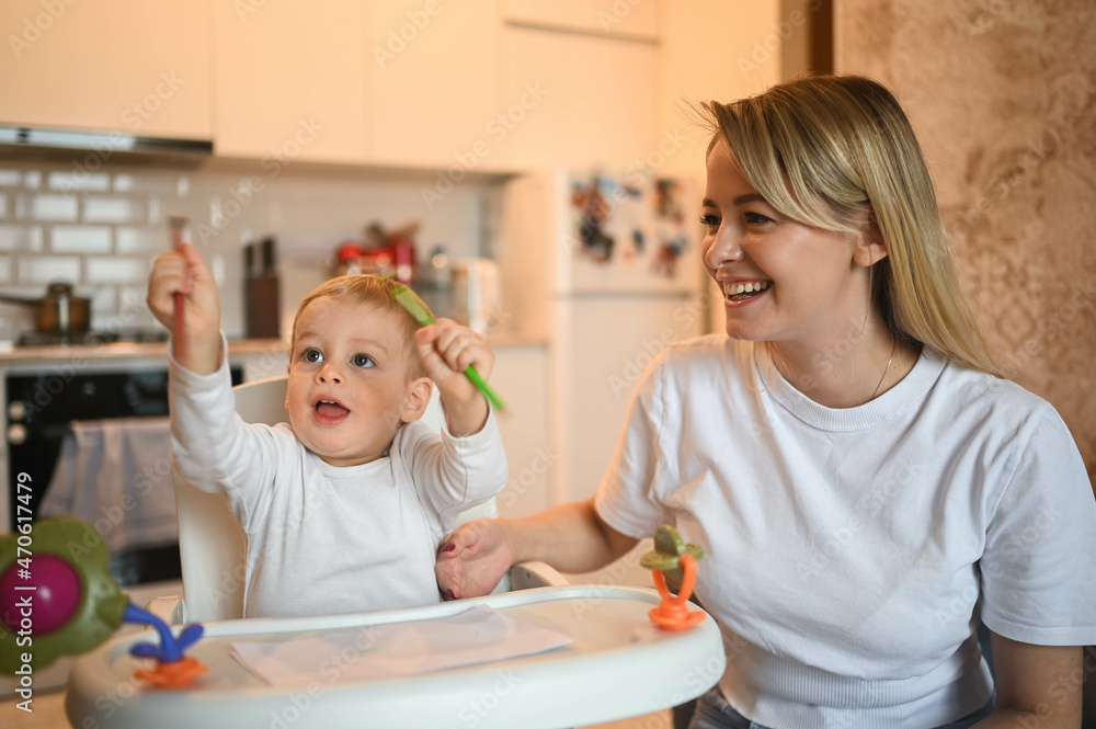Little cute baby toddler boy blonde sitting on baby chair learning to draw. Beautiful young mom and son playing spend time together indoors at home with toys. Healthy happy family childhood concept Little cute baby toddler boy blonde sitting on baby chair learning to draw. Beautiful young mom and son playing spend time together indoors at home with toys. Healthy happy family childhood concept