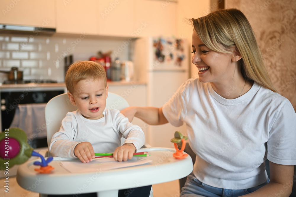 Little cute baby toddler boy blonde sitting on baby chair learning to draw. Beautiful young mom and son playing spend time together indoors at home with toys. Healthy happy family childhood concept