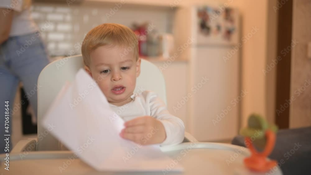 Little cute baby toddler boy blonde sitting on baby chair learning draw with colour pencils. Baby facial expressions indoors at home kitchen interior with toys. Healthy happy family childhood concept Little cute baby toddler boy blonde sitting on baby chair learning draw with colour pencils. Baby facial expressions indoors at home kitchen interior with toys. Healthy happy family childhood concept