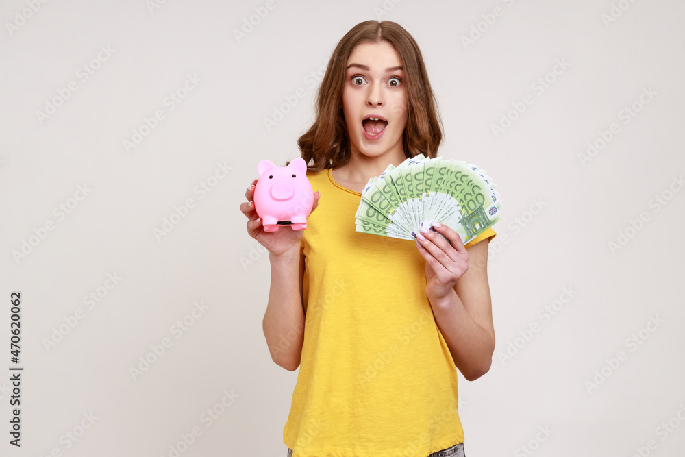 Extremely shocked female of young age in yellow T-shirt holding piggy bank and euro banknotes looking at camera with open mouth, deposit, cashback. Indoor studio shot isolated on gray background. Extremely shocked female of young age in yellow T-shirt holding piggy bank and euro banknotes looking at camera with open mouth, deposit, cashback. Indoor studio shot isolated on gray background.