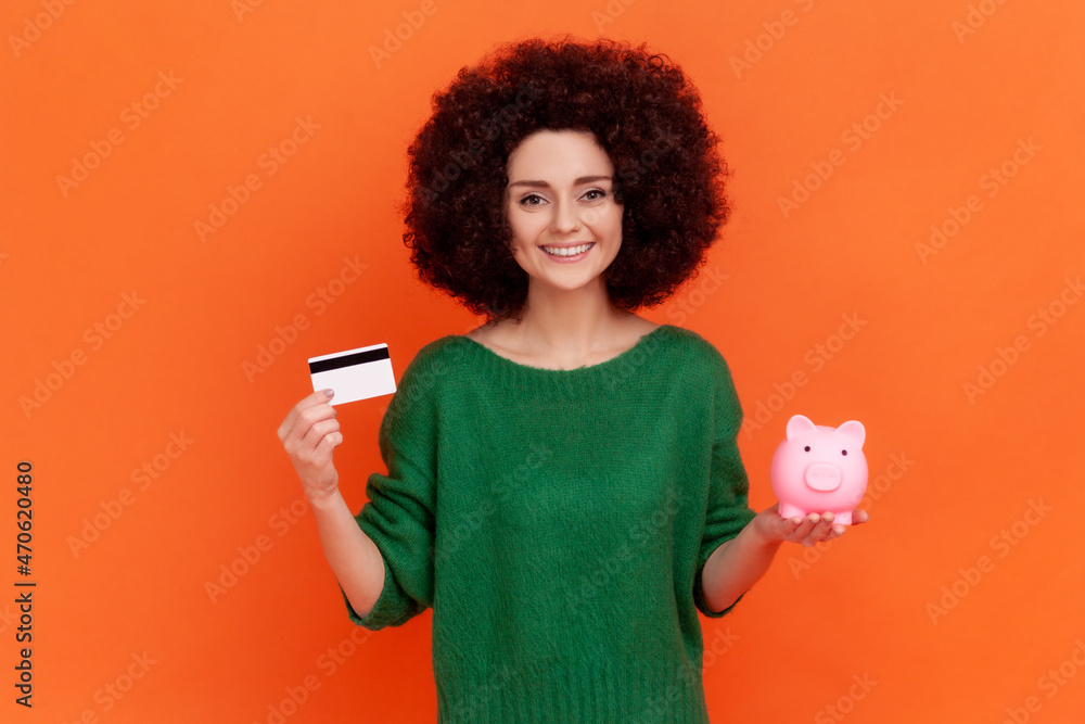 Φωτογραφία Positive kind young adult woman with Afro hairstyle wearing green casual style sweater standing showing to camera piggy bank and credit card