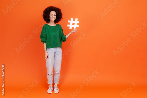 Full length portrait of woman with Afro hairstyle wearing green casual style sweater presenting white hashtag symbol, internet popularity. Indoor studio shot isolated on orange background. photo