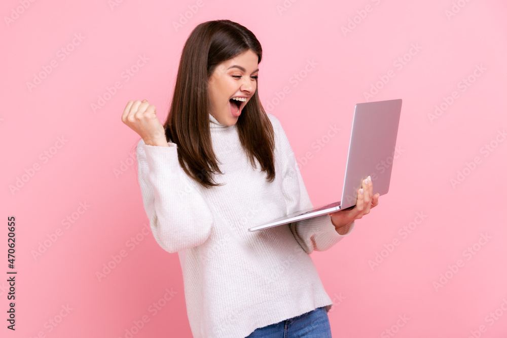 Satisfied female standing with laptop and clenching her fist, celebrating success, yelling happily, wearing white casual style sweater. Indoor studio shot isolated on pink background.