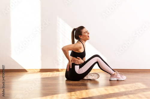 Side view of woman touching her painful back, being injured during sport exercising, wearing black sports top and tights. Full length studio shot illuminated by sunlight from window.