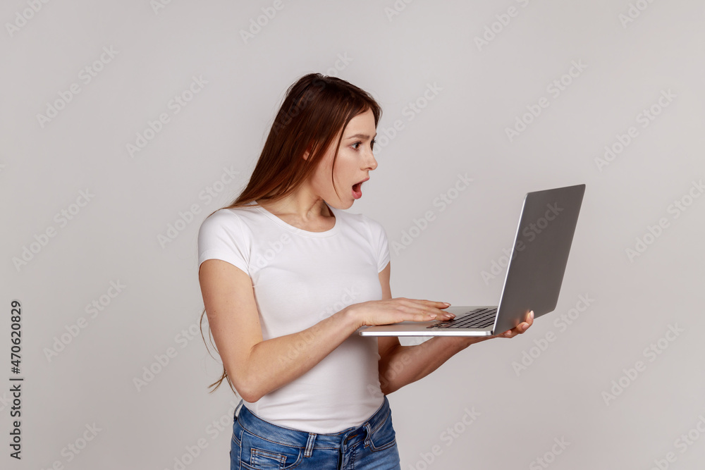 Portrait of astonished woman with dark hair working online, looking at laptop display with shocked expression, wearing white T-shirt. Indoor studio shot isolated on gray background. Portrait of astonished woman with dark hair working online, looking at laptop display with shocked expression, wearing white T-shirt. Indoor studio shot isolated on gray background.