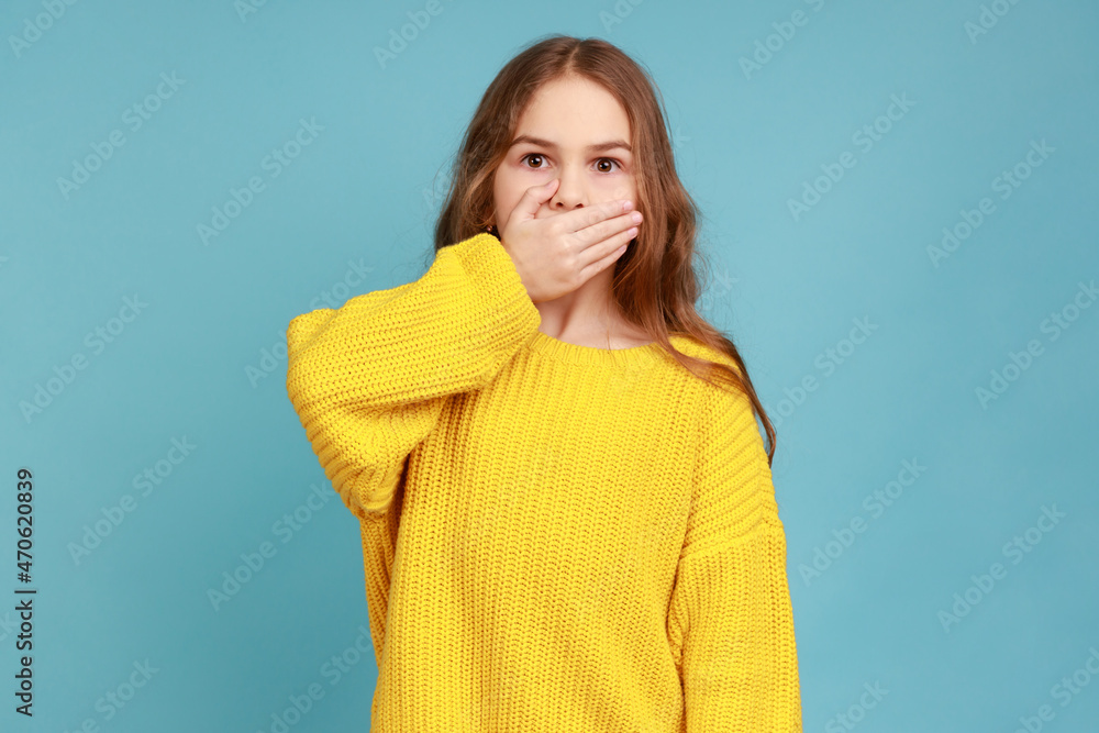 Portrait of little girl covering mouth with hand, afraid to say secret, child terrified to speak, wearing yellow casual style sweater. Indoor studio shot isolated on blue background. Portrait of little girl covering mouth with hand, afraid to say secret, child terrified to speak, wearing yellow casual style sweater. Indoor studio shot isolated on blue background.