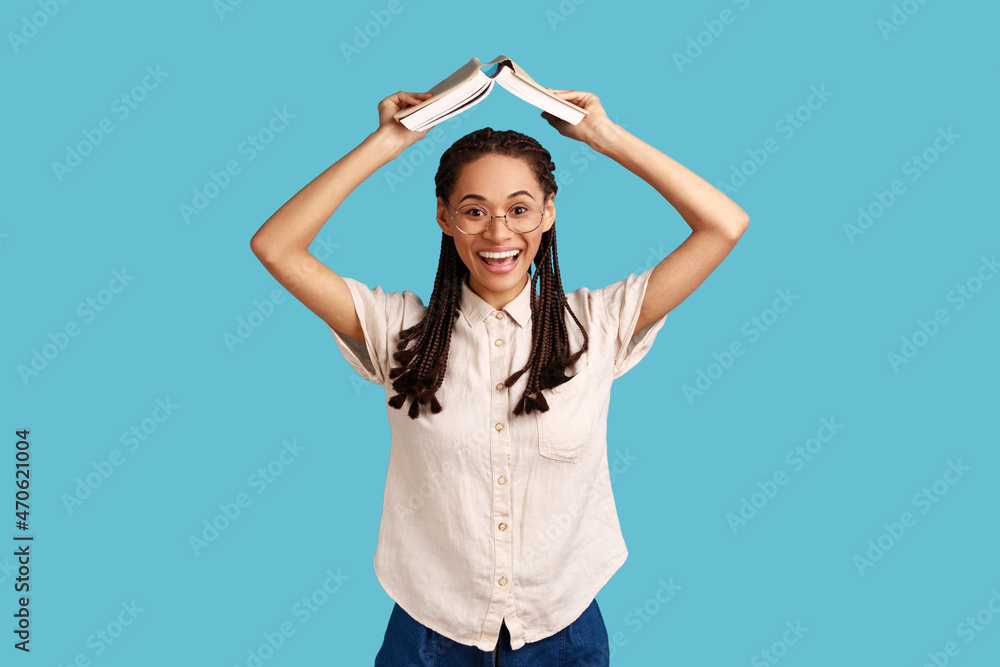 Portrait of positive woman with black dreadlocks and eyeglasses, standing holding book over head, studying and education, wearing white shirt. Indoor studio shot isolated on blue background. Portrait of positive woman with black dreadlocks and eyeglasses, standing holding book over head, studying and education, wearing white shirt. Indoor studio shot isolated on blue background.