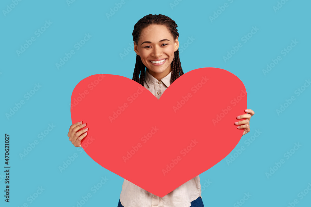 Portrait of attractive smiling woman with black dreadlocks holding big red heart, expressing positive romantic emotions, wearing white shirt. Indoor studio shot isolated on blue background. Portrait of attractive smiling woman with black dreadlocks holding big red heart, expressing positive romantic emotions, wearing white shirt. Indoor studio shot isolated on blue background.