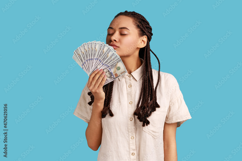 Attractive rich woman with black dreadlocks holding big fan of money, smelling dollar banknotes with pleasure, wearing white shirt. Indoor studio shot isolated on blue background. Attractive rich woman with black dreadlocks holding big fan of money, smelling dollar banknotes with pleasure, wearing white shirt. Indoor studio shot isolated on blue background.