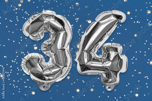 Silver foil balloon number, digit thirty four on a blue background with sequins. Birthday greeting card with inscription 34. Top view. Numerical digit. Celebration event, template. photo