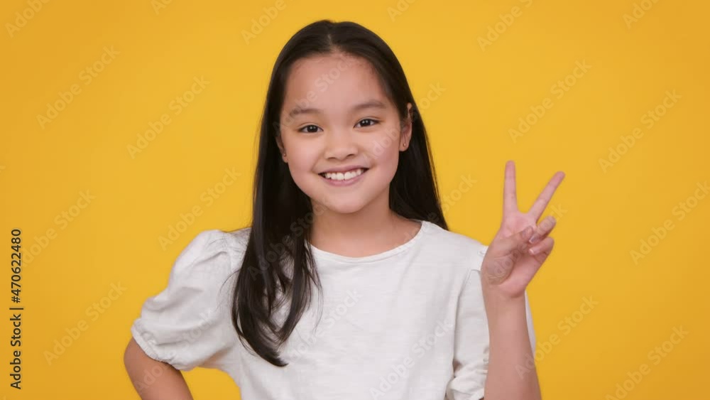 Happy childhood. Adorable preteen asian girl model gesturing V sign and smiling to camera, orange studio background Stock ビデオ | Adobe Stock