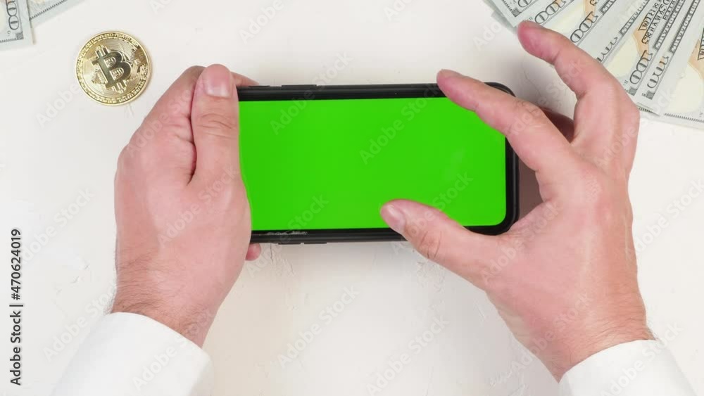 Hands of a successful young financier in a shirt hold a smart phone with a chroma key mockup for application on a white background with metallic bitcoin and dollars Hands of a successful young financier in a shirt hold a smart phone with a chroma key mockup for application on a white background with metallic bitcoin and dollars