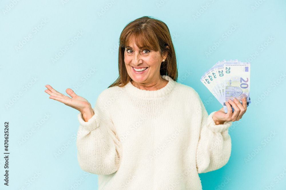 Obraz premium Middle age caucasian woman holding bank notes isolated on blue background showing a copy space on a palm and holding another hand on waist.