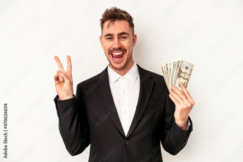 Young caucasian business man holding bank notes isolated on white background joyful and carefree showing a peace symbol with fingers. Young caucasian business man holding bank notes isolated on white background joyful and carefree showing a peace symbol with fingers.