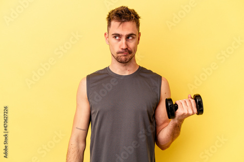 Young sport man holding a dumbbell isolated on yellow background confused, feels doubtful and unsure.