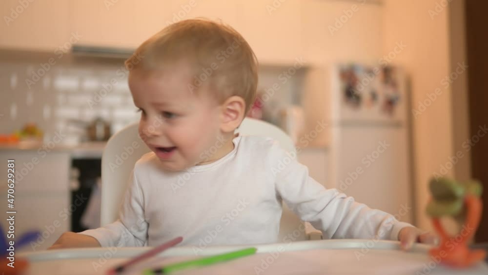 Little cute baby toddler boy blonde sitting on baby chair learning draw with colour pencils. Baby facial expressions indoors at home kitchen interior with toys. Healthy happy family childhood concept Little cute baby toddler boy blonde sitting on baby chair learning draw with colour pencils. Baby facial expressions indoors at home kitchen interior with toys. Healthy happy family childhood concept