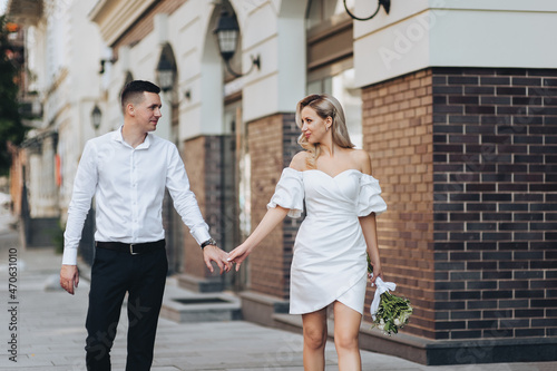 A smiling, stylish groom and a beautiful blonde bride in a white dress are walking holding hands along the street in the city against the background of ancient buildings. Wedding photography.