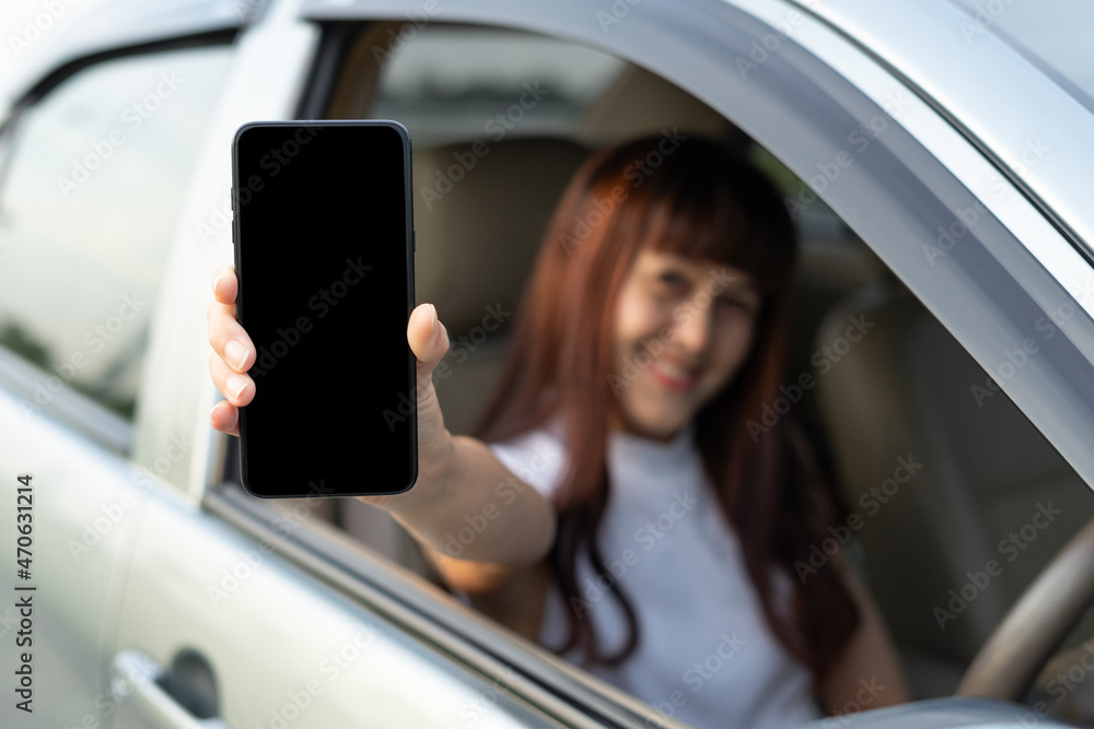 Happy female driver smiling and showing smartphone while through car window. View of a lady driving her car to travel on her holiday vacation time. Loan car finance insurance, buy and rental concept.
