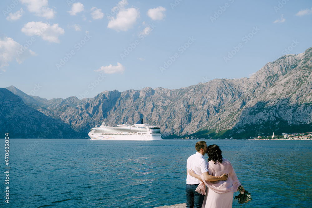 Bride and groom stand on the pier against the backdrop of the liner and mountains. Back view
