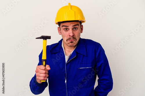 Young caucasian worker man holding a hammer isolated on white background shrugs shoulders and open eyes confused.