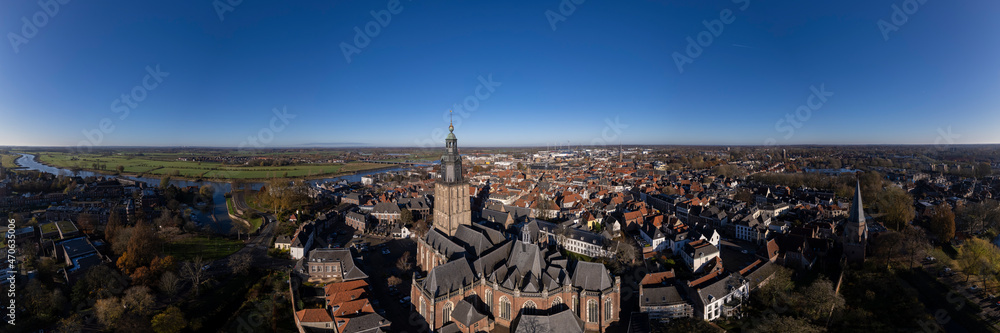 Tower town Zutphen in The Netherlands with medieval Hanseatic city center on the river IJssel and Walburgiskerk centrally in the frame. Aerial panorama of Dutch settlement against a clear blue sky.