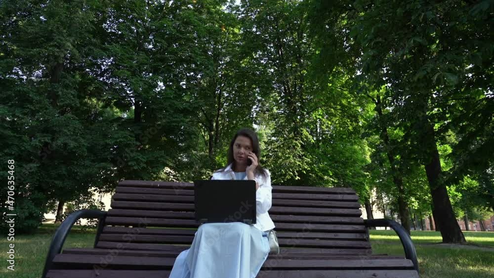 Adult woman talks on phone working on laptop in park sitting on bench and smiling in white jacket and long dress slow motion Adult woman talks on phone working on laptop in park sitting on bench and smiling in white jacket and long dress slow motion
