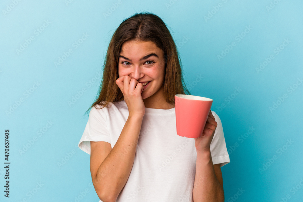 Young caucasian woman holding a mug isolated on blue background biting fingernails, nervous and very anxious. Young caucasian woman holding a mug isolated on blue background biting fingernails, nervous and very anxious.