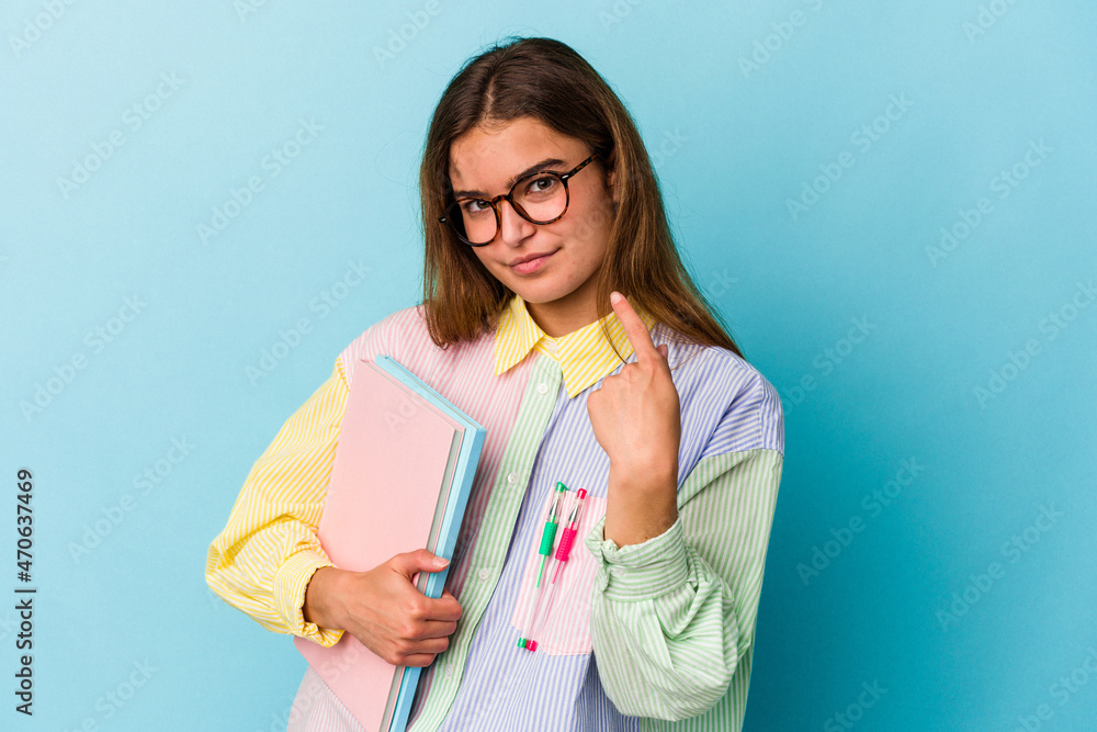 Young caucasian student woman holding books isolated on blue background pointing with finger at you as if inviting come closer. Young caucasian student woman holding books isolated on blue background pointing with finger at you as if inviting come closer.