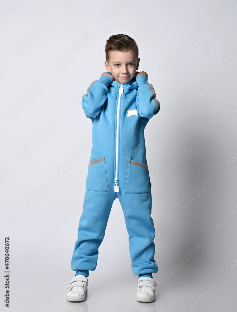 Naklejka premium Smiling kid boy in blue jumpsuit with zipper and reflective stripes and white sneakers stands holding his cowl with hands, putting on hood over light background