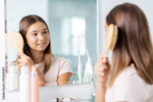 beauty and people concept - teenage girl looking to mirror and brushing hair with comb at home bathroom