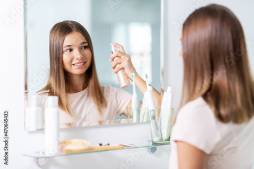 beauty and people concept - teenage girl looking to mirror and using hair styling spray at home bathroom