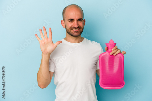 Young bald man holding a hot bag of water isolated on blue background smiling cheerful showing number five with fingers.