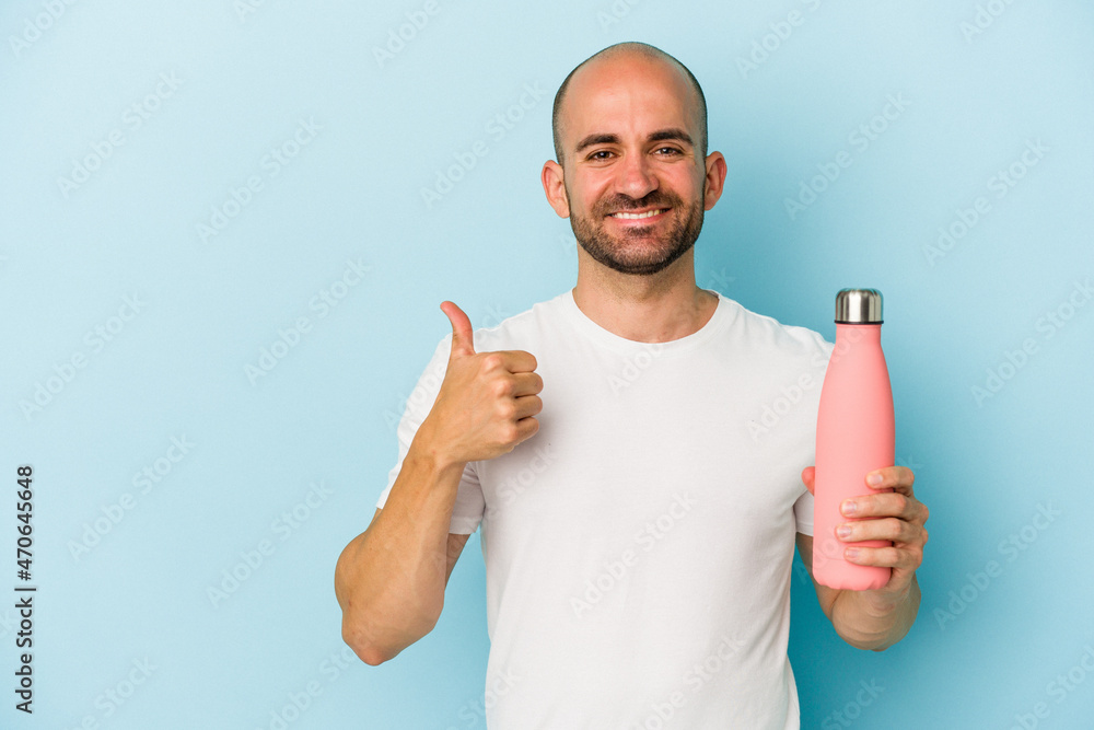 Young bald man holding canteen isolated on blue background smiling and raising thumb up Young bald man holding canteen isolated on blue background smiling and raising thumb up