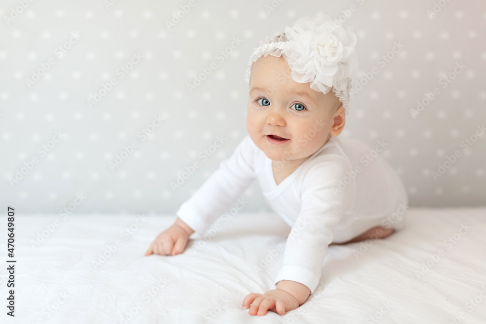 Happy toddler baby girl in a white bodysuit sitting on bed at home and looking at the camera with smile. Copy space for text Happy toddler baby girl in a white bodysuit sitting on bed at home and looking at the camera with smile. Copy space for text