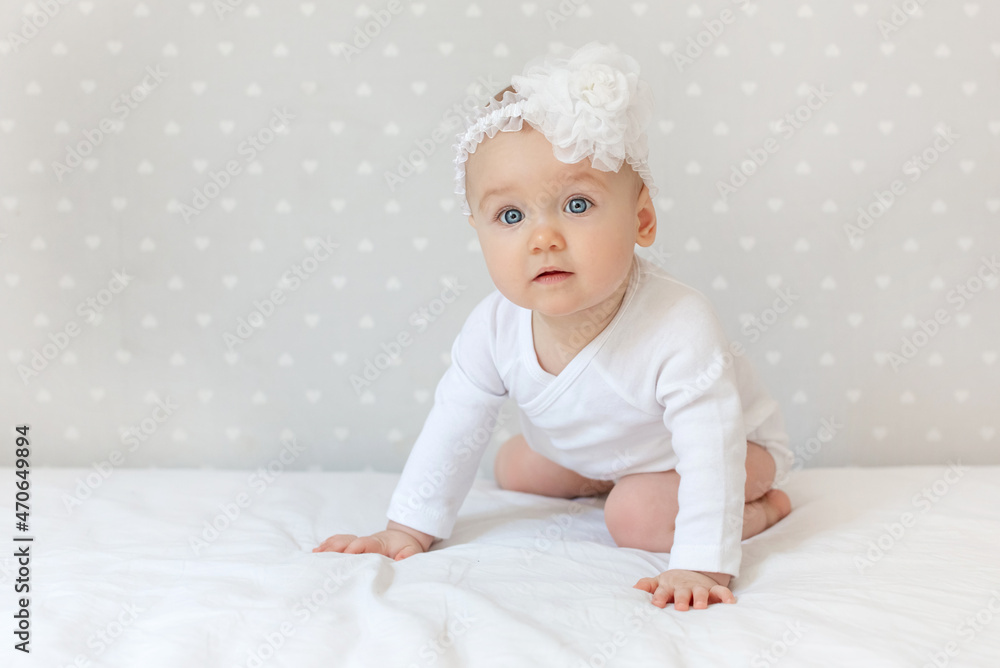 Happy toddler baby girl in a white bodysuit sitting on bed at home and looking at the camera with smile. Copy space for text Happy toddler baby girl in a white bodysuit sitting on bed at home and looking at the camera with smile. Copy space for text