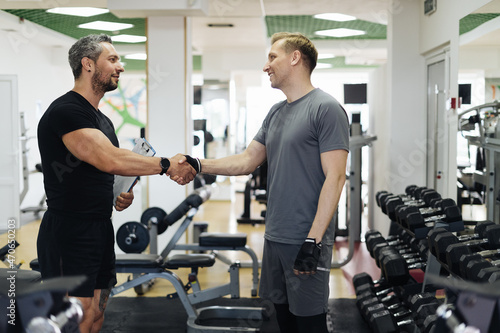 Fit man discussing workout plan with his trainer, saing hello before starting workout in the gym. Personal coach standing near young man and showing something on his clipboard