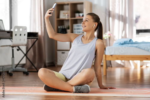 sport, fitness and healthy lifestyle concept - smiling teenage girl with smartphone and earphones sitting on yoga mat and taking selfie at home