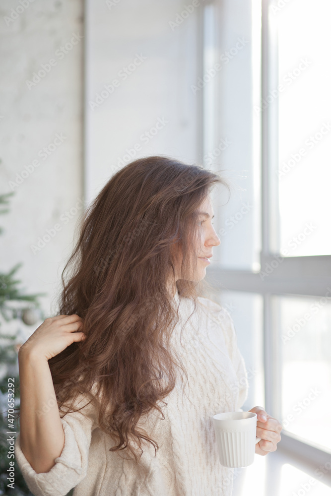 Young beautiful woman holding a cup of coffee or tea standing by the window and christmas tree Young beautiful woman holding a cup of coffee or tea standing by the window and christmas tree