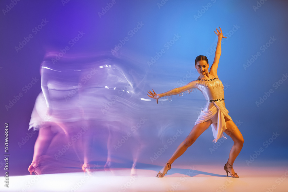 Portrait of young graceful flexible woman dancing ballroom dance without partner isolated on gradient blue purple background in neon mixed light
