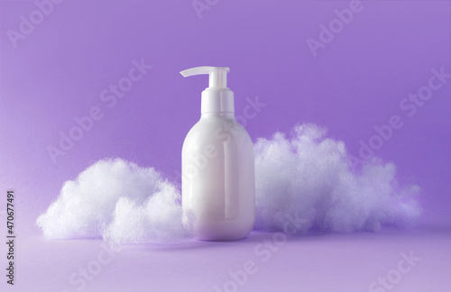 Lotion with fluffy clouds on violet background. Cosmetic white plastic bottle with pump beauty product for skincare or body care. Concept care with sensitive skin. Creative still-life photo